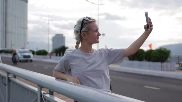 Красивая стройная женщина с косичкой-блондинкой в серой футболке, стоящая на мосту и делающая селфи на мобильном телефоне на фоне города. Девчачий экран — стоковое видео