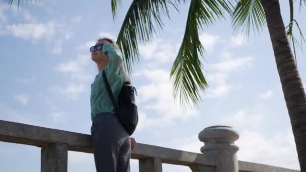 Heureuse femme caucasienne avec de longs cheveux blonds en lunettes de soleil et chemise verte debout et souriante près du palmier sur un fond bleu ciel. Concept de voyage — Video