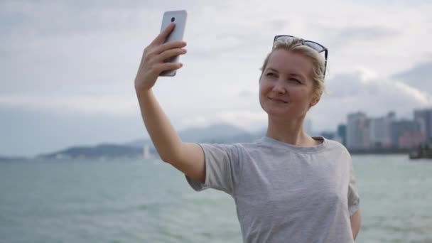 Hermosa mujer delgada con una rubia pigtail en una camiseta gris de pie en el puente y haciendo selfie en el teléfono móvil sobre el fondo del mar y la ciudad. Chica tocando pantalla — Vídeos de Stock