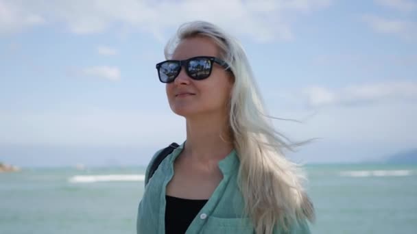 Jeune femme caucasienne heureuse avec de longs cheveux blonds en lunettes de soleil et chemise verte debout et souriante près du palmier sur un fond bleu de mer. Concept de voyage — Video