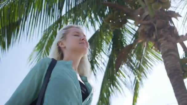 Joven mujer caucásica feliz con el pelo largo y rubio en gafas de sol y camisa verde de pie con los ojos cerrados y sonriendo cerca de la palmera sobre un fondo azul cielo. Concepto de viaje — Vídeo de stock