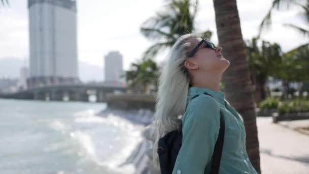 Heureuse femme caucasienne aux longs cheveux blonds en lunettes de soleil et chemise verte debout et souriante près d'un palmier sur fond de parc et de mer. Concept de voyage — Video