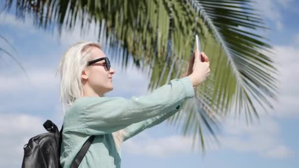 Belle femme mince avec de longs cheveux blonds en lunettes de soleil et chemise verte debout près de palmier et prendre des photos par smartphone sur un fond bleu ciel . — Video