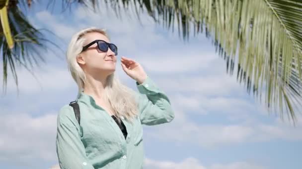 Mulher caucasiana feliz com longos cabelos loiros em óculos de sol e camisa verde de pé e sorrindo perto de palmeira em um fundo azul céu. Conceito de viagem — Vídeo de Stock