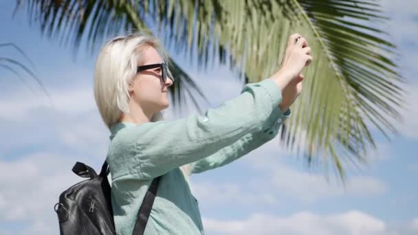 Palmiye ağacında yakın güneş gözlüğü ve yeşil gömlek ayakta uzun sarışın saçlı güzel ince kadın ve mavi gökyüzü arka planda akıllı telefon tarafından bir fotoğraf çekmek. — Stok video