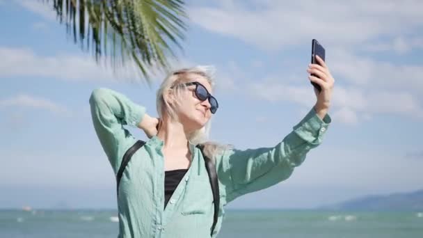 Piękna, szczupła kobieta z długie blond włosy, Okulary przeciwsłoneczne i zielonej koszuli stojący w pobliżu palmy i podejmowania selfie na telefon komórkowy na niebieskim tle nieba i morza. Dziewczyna przy użyciu smartfona — Wideo stockowe