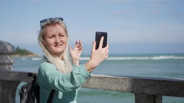 Belle femme mince avec de longs cheveux blonds en lunettes de soleil et chemise verte debout près de palmier et faire du selfie sur un téléphone portable sur un ciel bleu et fond de mer. Fille à l'aide smartphone — Video