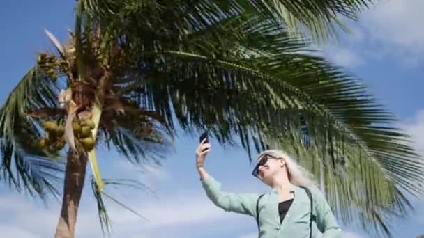 Mulher fina bonita com cabelo loiro longo em óculos de sol e camisa verde de pé perto da palmeira e fazendo selfie no telefone celular em um céu azul e fundo do mar. Menina usando smartphone — Vídeo de Stock