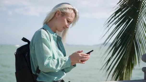Hermosa mujer delgada con el pelo largo y rubio y camisa verde de pie cerca de la palmera y el uso de teléfono inteligente sobre el fondo del mar. Chica tocando pantalla — Vídeos de Stock