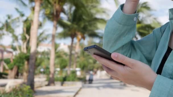 Palmiye ağacında yakın yeşil gömlek ayakta uzun sarı saçlı güzel ince kadın ve arka planda Park üzerinden akıllı telefon kullanarak. Kare dokunmadan ekran ve gülümseme kız. — Stok video