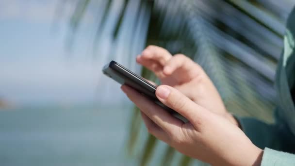 Mãos femininas usando smartphone sobre o fundo do mar e palmeiras. Menina tocando tela — Vídeo de Stock
