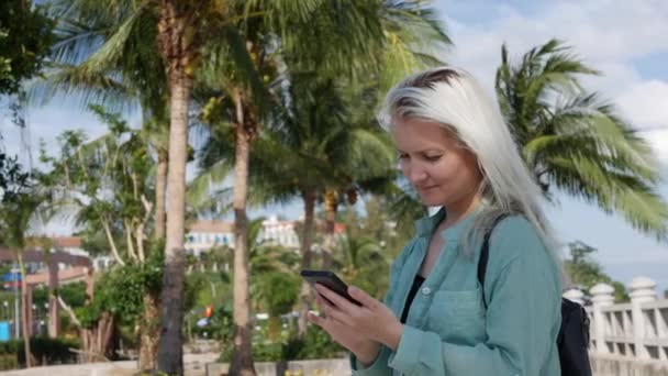 Schöne schlanke Frau mit langen blonden Haaren in grünem Hemd, die in der Nähe von Palmen steht und über dem Hintergrund des Parks ihr Smartphone benutzt. Mädchen auf dem Platz berührt Bildschirm und lächelt. — Stockvideo