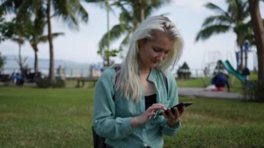 Yeşil gömlek uzun sarışın saçlı güzel ince kadın yere oturur ve arka planda Park üzerinden akıllı telefon kullanarak. Kare dokunmadan ekran ve gülümseme kız.