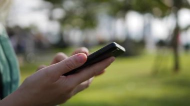 Yeşil gömlek uzun sarışın saçlı Genç güzel ince kadın yere oturur ve arka planda Park üzerinden akıllı telefon kullanarak. Kare dokunmadan ekran ve gülümseme kız.