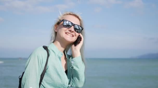 Beautiful slim woman with long blonde hair and green shirt standing and talking on phone over background sea. — Stock Video