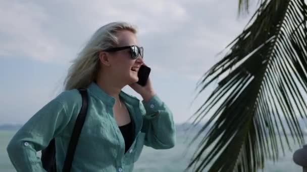 Mujer caucásica feliz con el pelo largo y rubio en gafas de sol y camisa verde de pie y hablando por teléfono cerca de la palmera en un cielo azul y fondo marino. Concepto de viaje — Vídeos de Stock