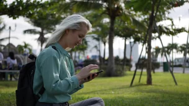 Mulher magro bonita com cabelo loiro longo em camisa verde senta-se no chão e usando smartphone sobre o fundo do parque. Menina na tela de toque quadrado e sorriso . — Vídeo de Stock