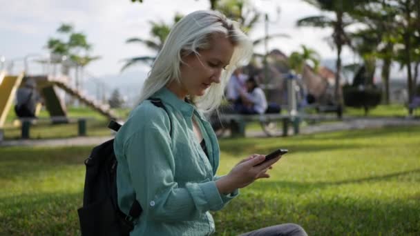 Belle femme mince avec de longs cheveux blonds en chemise verte est assis sur le sol et en utilisant smartphone sur fond le parc. Fille sur l'écran tactile carré et sourire . — Video