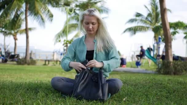 Piękna szczupła kobieta z długimi blond włosami w zielonej koszulce siedzi na ziemi i za pomocą smartfona nad tłem parku. Dziewczyna na placu dotykając ekranu i uśmiech. — Wideo stockowe