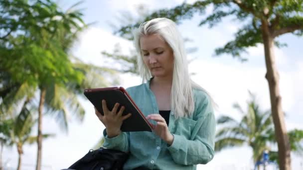 Piękna szczupła kobieta z długimi blond włosami w zielonej koszulce siedzi na ziemi i za pomocą smartfona nad tłem parku. Dziewczyna na placu dotykając ekranu i uśmiech. — Wideo stockowe
