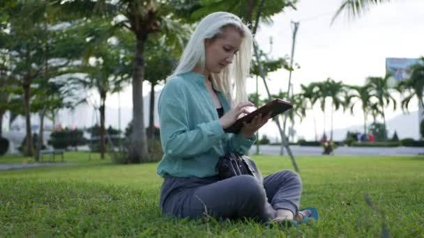 Piękna szczupła kobieta z długimi blond włosami w zielonej koszulce siedzi na ziemi i za pomocą smartfona nad tłem parku. Dziewczyna na placu dotykając ekranu i uśmiech. — Wideo stockowe