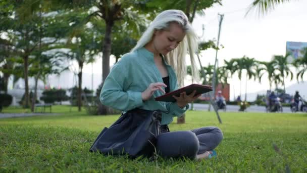 Mulher magro bonita com cabelo loiro longo em camisa verde senta-se no chão e usando smartphone sobre o fundo do parque. Menina na tela de toque quadrado e sorriso . — Vídeo de Stock