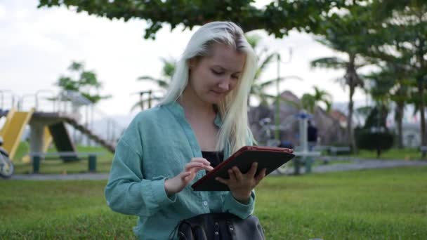 Belle femme mince avec de longs cheveux blonds en chemise verte est assis sur le sol et en utilisant smartphone sur fond le parc. Fille sur l'écran tactile carré et sourire . — Video