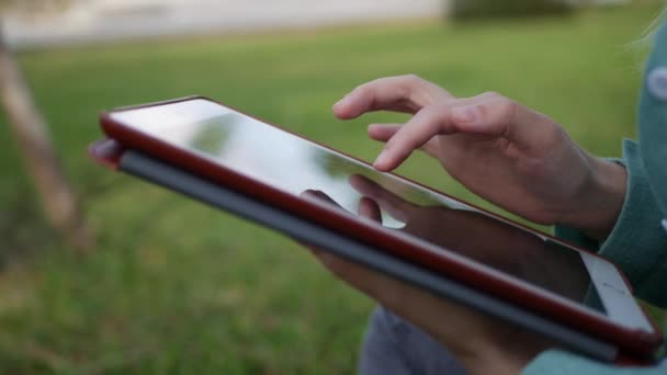 Ung vacker smal kvinna med långt blont hår i grön skjorta sitter på marken och använder Tablet PC över bakgrunden parken. Flicka på torget röra skärmen och leende. — Stockvideo