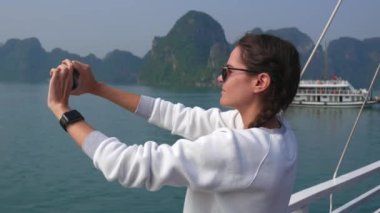 Akıllı Saat beyaz bir kazak ve güneş gözlüğü takan genç kadın bir smartphone bir teknede dururken körfezde resmini yapar. Seyahat kavramı