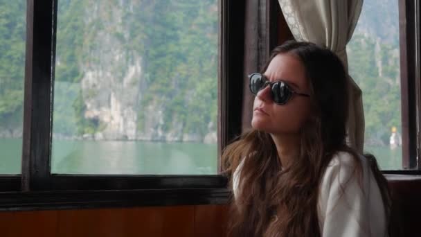 Een jonge vrouw zit in een boot in een witte trui en zonnebril kijkt de prachtige uitzicht op de baai. Travel concept — Stockvideo