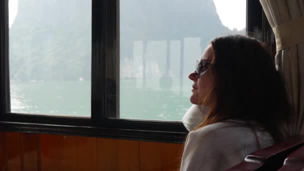 Młoda kobieta siedzi wewnątrz łodzi w biały sweter i okulary patrzy się na piękny widok na zatokę. Koncepcja podróży — Wideo stockowe