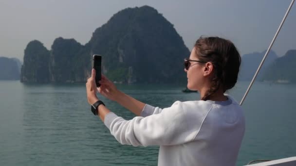 Mujer joven con reloj inteligente en un suéter blanco y gafas de sol hace la imagen de la bahía en un teléfono inteligente mientras está de pie en un barco. Concepto de viaje — Vídeo de stock