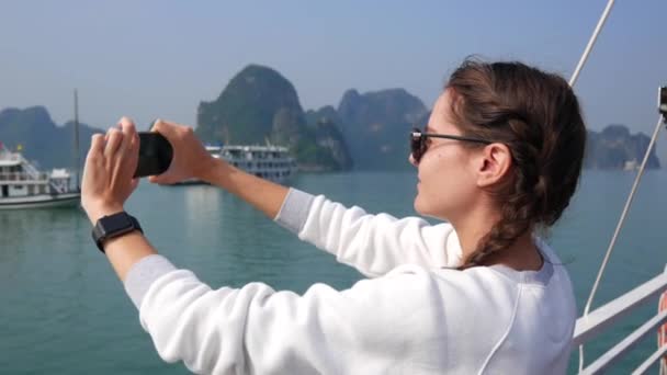 Mujer joven con reloj inteligente en un suéter blanco y gafas de sol hace la imagen de la bahía en un teléfono inteligente mientras está de pie en un barco. Concepto de viaje — Vídeo de stock