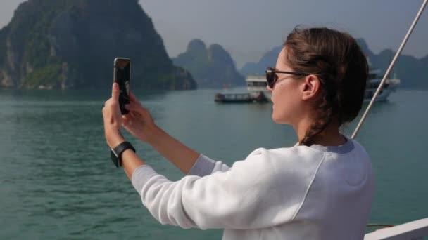 Mujer joven con reloj inteligente en un suéter blanco y gafas de sol hace la imagen de la bahía en un teléfono inteligente mientras está de pie en un barco. Concepto de viaje — Vídeos de Stock