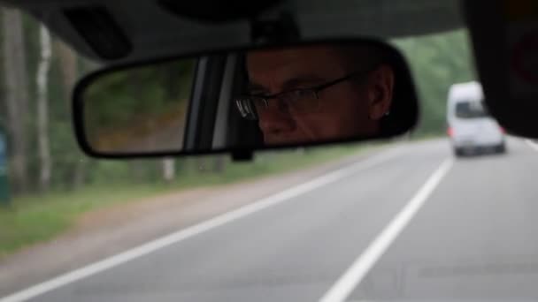 Un hombre mayor conduce un coche en la carretera del campo. Reflejo de la cara en el espejo retrovisor del auto. Primer plano. 4k — Vídeo de stock