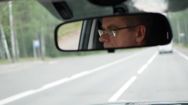 Un hombre mayor conduce un coche en la carretera del campo. Reflejo de la cara en el espejo retrovisor del auto. Primer plano. — Vídeos de Stock