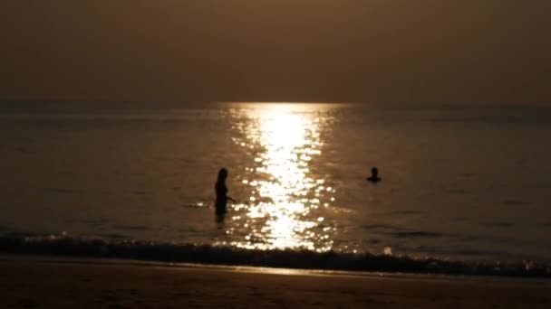 Verschwommene Silhouetten von Menschen, die am Strand spazieren und bei Sonnenuntergang im Meer schwimmen. Ferienkonzept. Nahaufnahme. 4k — Stockvideo