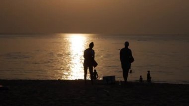 Sahilde yürüyen ve gün batımında denizde yüzen insanların siluetleri. Tatil konsepti. Closeup. 4k
