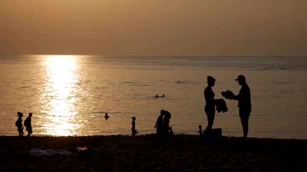 Silhouetten von Menschen, die am Strand spazieren und bei Sonnenuntergang im Meer schwimmen. Ferienkonzept. Nahaufnahme. 4k — Stockvideo