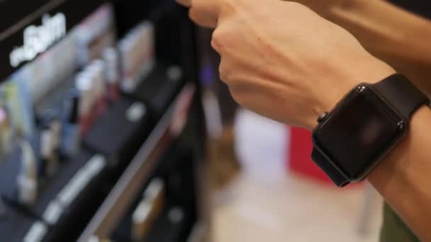 Jovem com um relógio inteligente seleciona lápis de sobrancelha e testes em sua mão no centro comercial. Mesa com cosméticos, batom, sombras, tinta para pestanas, espelho, pincéis de maquiagem. Fecha a porta. 4k — Vídeo de Stock