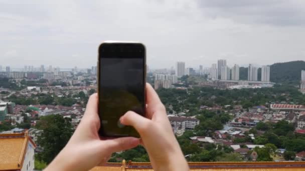 Young woman makes vertical photos of city on the smartphone standing on the mountain. Camera over the shoulder. Cloudy sky. Closeup. 4k — Stock Video