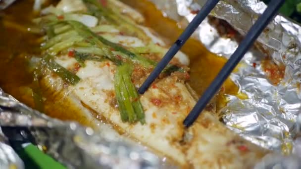 Os turistas comem peixe por pauzinhos cozidos em papel alumínio no mercado asiático de comida de rua. Fechar. — Vídeo de Stock