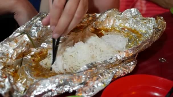 Os turistas comem macarrão de arroz com peixe cozido em papel alumínio no mercado asiático de comida de rua. Fechar — Vídeo de Stock