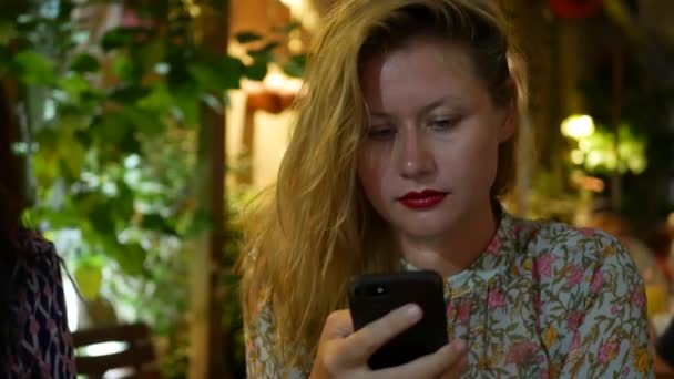 Pensive girl uses a smartphone, writes a message, chatting. Woman looking for information in the phone and sad. Sitting in restaurant alone. Evening time, lowlight. Technology and entertainment — Stock Video