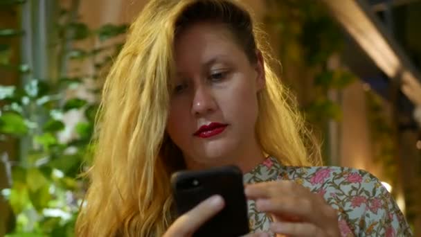 Pensive meisje maakt gebruik van een smartphone, schrijft een bericht, chatten. Vrouw op zoek naar informatie in de telefoon en verdrietig. Zittend in restaurant alleen. Avond tijd, lowlight. Technologie en entertainment — Stockvideo