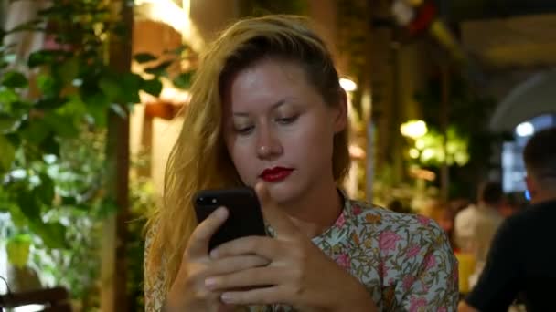 Pensive lány használ egy okostelefon, ír egy üzenetet, csevegni. Nő keresett információt a telefon és szomorú. Ül az étteremben egyedül. Esti idő, lowlight. Technológia-és szórakoztatóipar — Stock videók