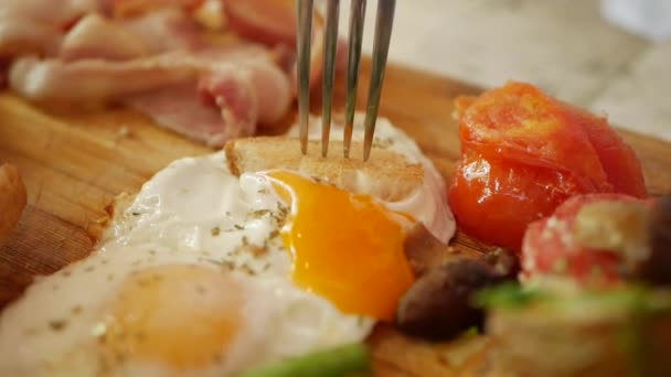 女性は、揚げ卵、ソーセージ、緑豆、ジャガイモ、キノコ、トマト、トースト、ハム、ベーコンと木製のボードで伝統的な英語の朝食を食べます。クローズ アップ — ストック動画