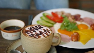Bir fincan latte ve geleneksel İngiliz kahvaltısı kızarmış yumurta, sosis, domates, jambon ve ahşap bir masada pastırma. Yakın plan.