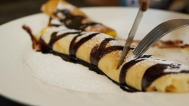 Çatal ve bıçak kullanan müşteri taze mango, çikolata ve pudra şekerli lezzetli krepler yiyor. Restoranda beyaz tabağa konmuş. Yakın plan.