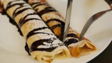 Çatal ve bıçak kullanan müşteri taze mango, çikolata ve pudra şekerli lezzetli krepler yiyor. Restoranda beyaz tabağa konmuş. Yakın plan.